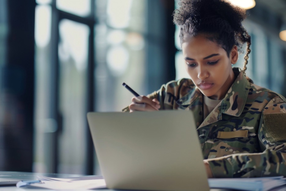Female military person sitting at a desk on a computer with dark curly hair and dark completed skin holding a pen in her right hand